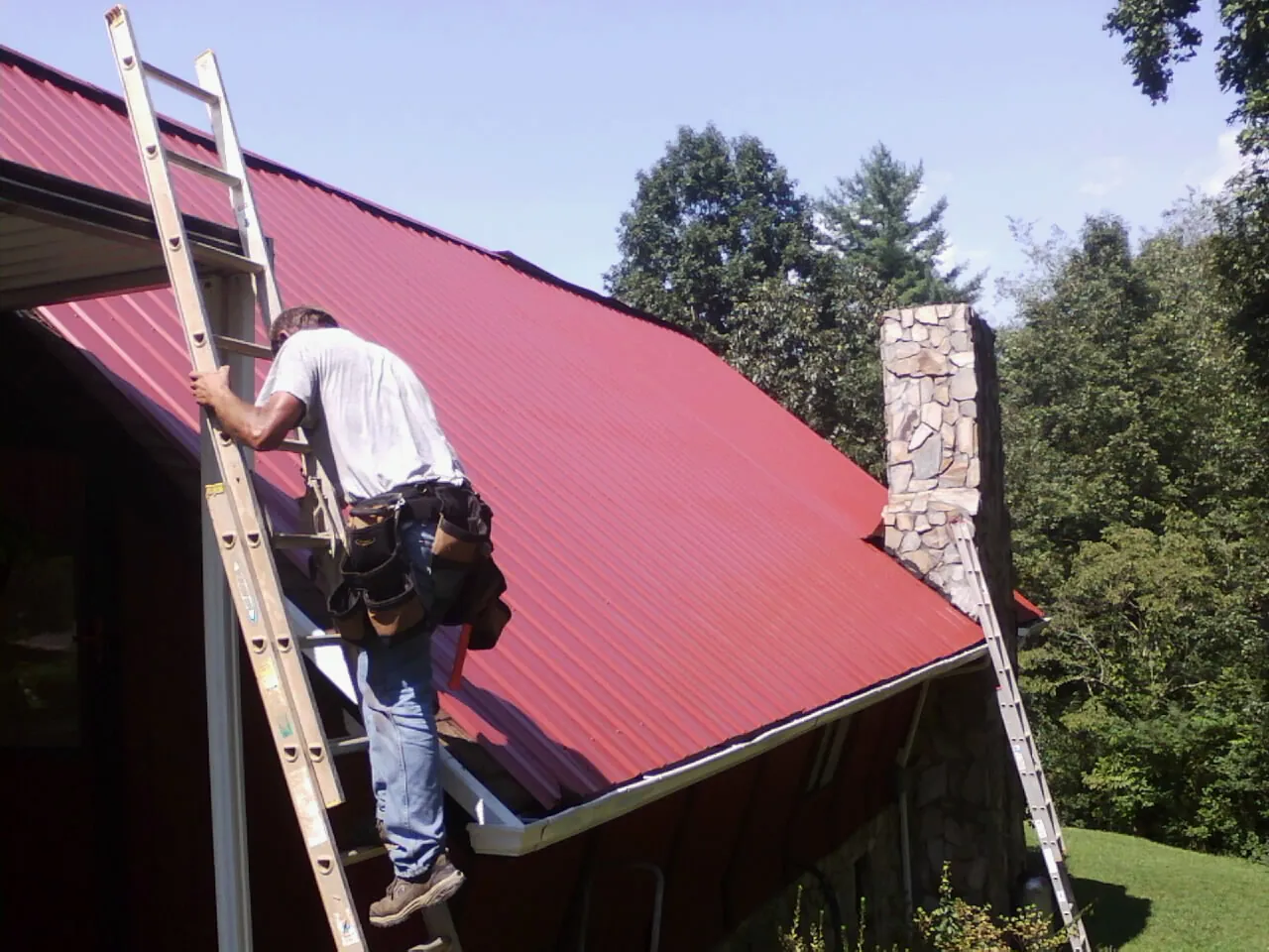 Roofer on a ladder beside a red metal roof and stone chimney — Soffit & Fascia Repair by Roofing Brothers in Polkville